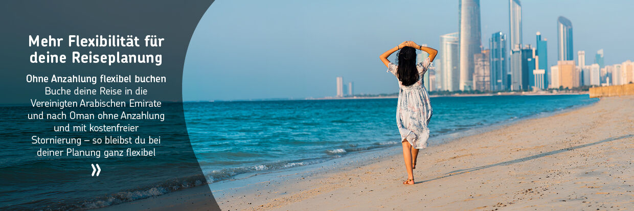 Frau in weißem Kleid mit Blumenmuster geht barfuß am Strand mit Blick auf eine moderne Stadt mit hohen Gebäuden