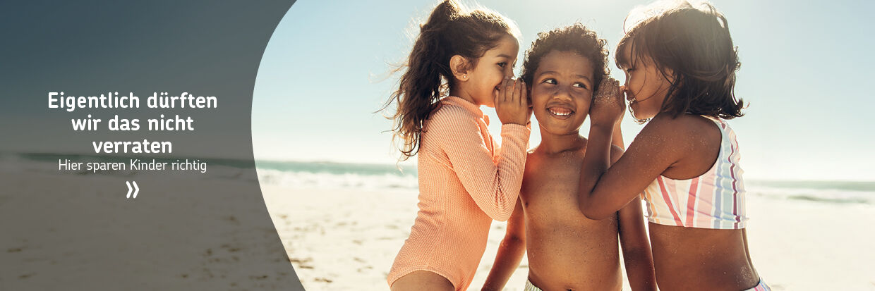 Drei Kinder in Badebekleidung am Strand, zwei flüstern dem dritten ins Ohr, alle lächeln