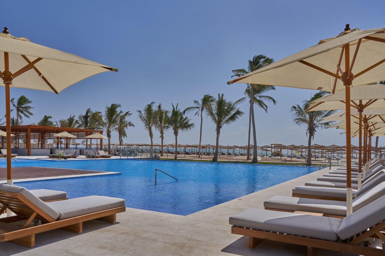 Großer Pool mit Liegen und Sonnenschirmen, im Hintergrund Palmen