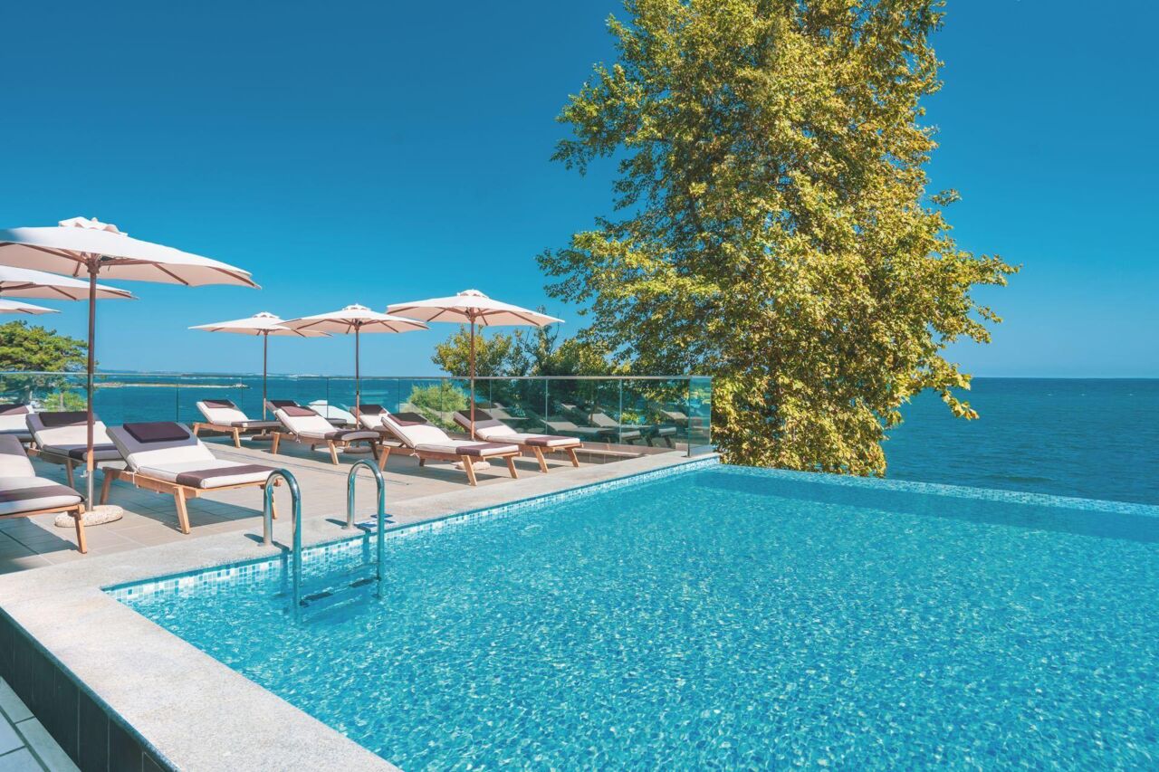Infinity-Pool mit Glasgeländer, Liegestühlen und Sonnenschirmen neben einem großen Baum mit Blick auf das Meer