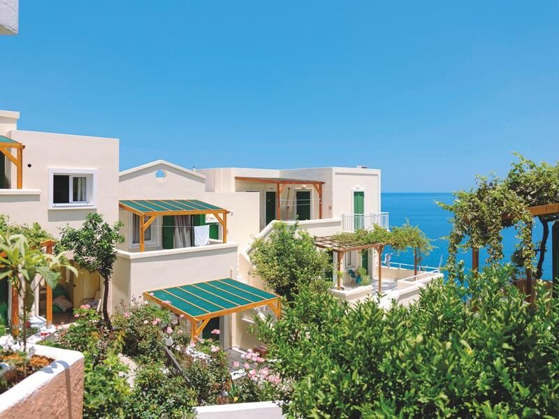 Mehrere weiße Häuser mit grünen Fensterläden und grünen Markisen vor blauem Himmel und Meer im Hintergrund