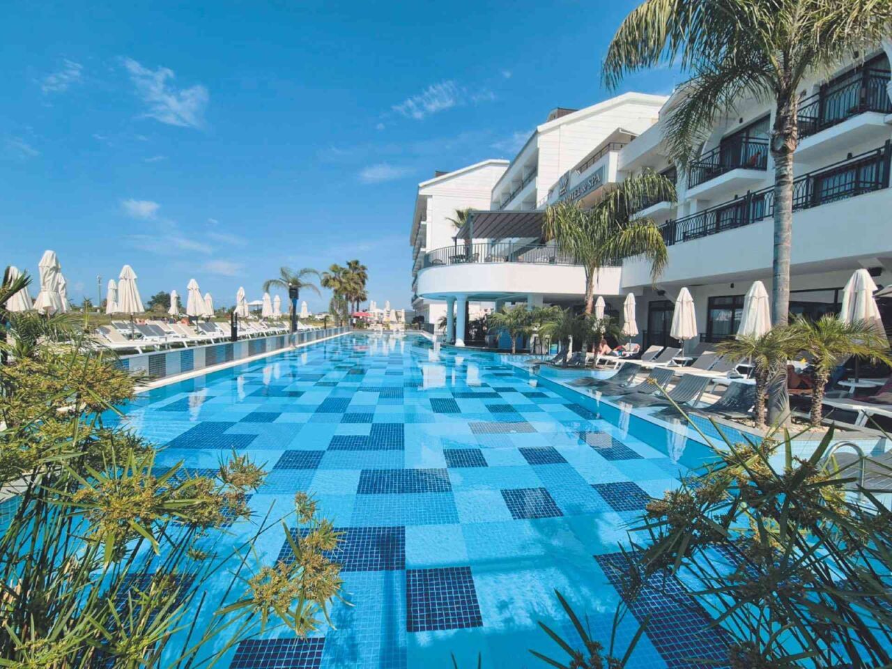 Großer Außenpool mit blau gemustertem Boden vor einem mehrstöckigen Hotel mit Balkonen und Palmen unter blauem Himmel