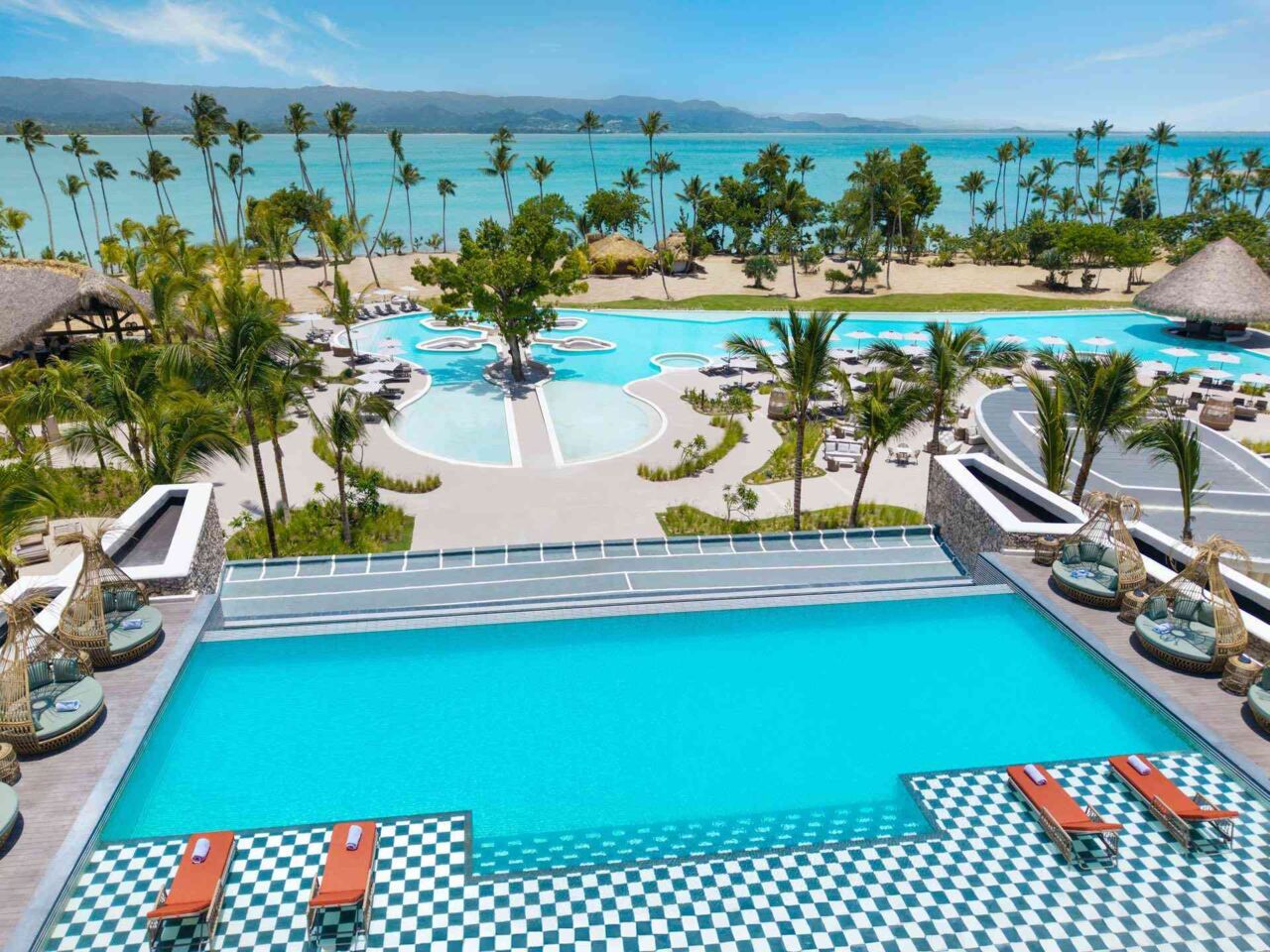 Großer rechteckiger Pool mit Liegen und Korbsesseln auf gemustertem Boden vor tropischem Garten mit Palmen und Strand am Meer