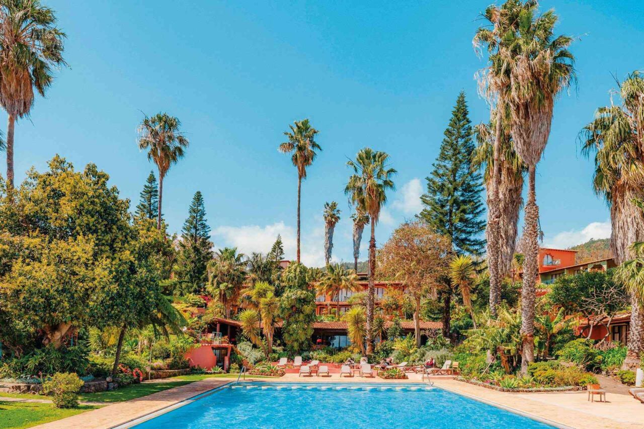 Großer Außenpool vor einem von Palmen und anderen Bäumen umgebenen mehrstöckigen Gebäude