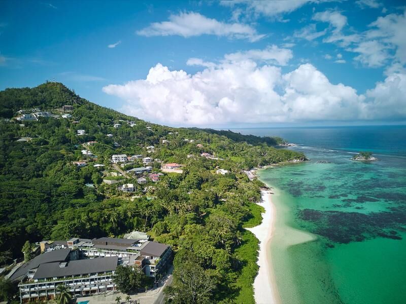 Luftaufnahme einer grünen Küstenlandschaft mit Gebäuden am Hang und einem weißen Sandstrand