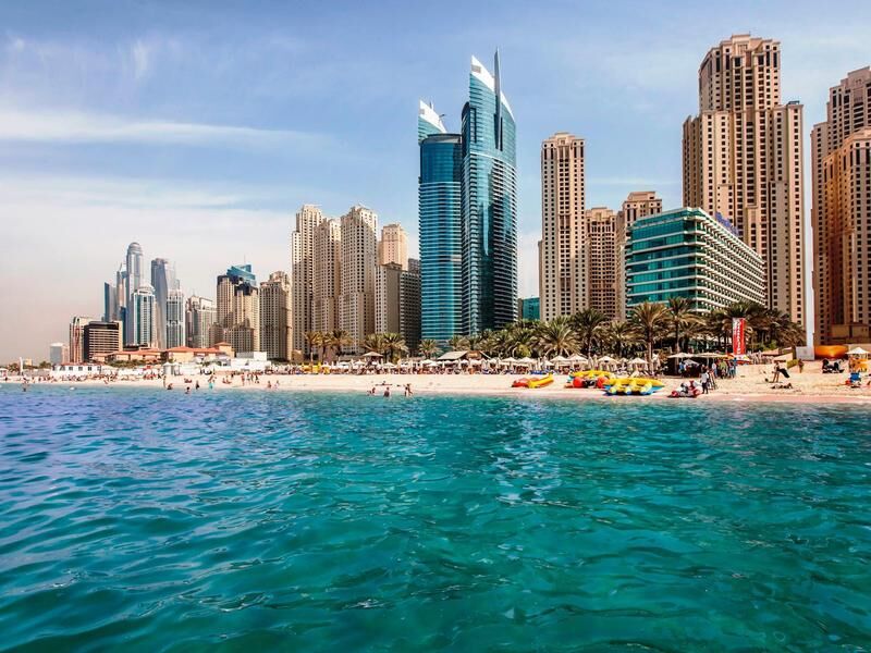 Strand mit Menschen und Palmen vor moderner Hochhaus-Skyline bei klarem Himmel