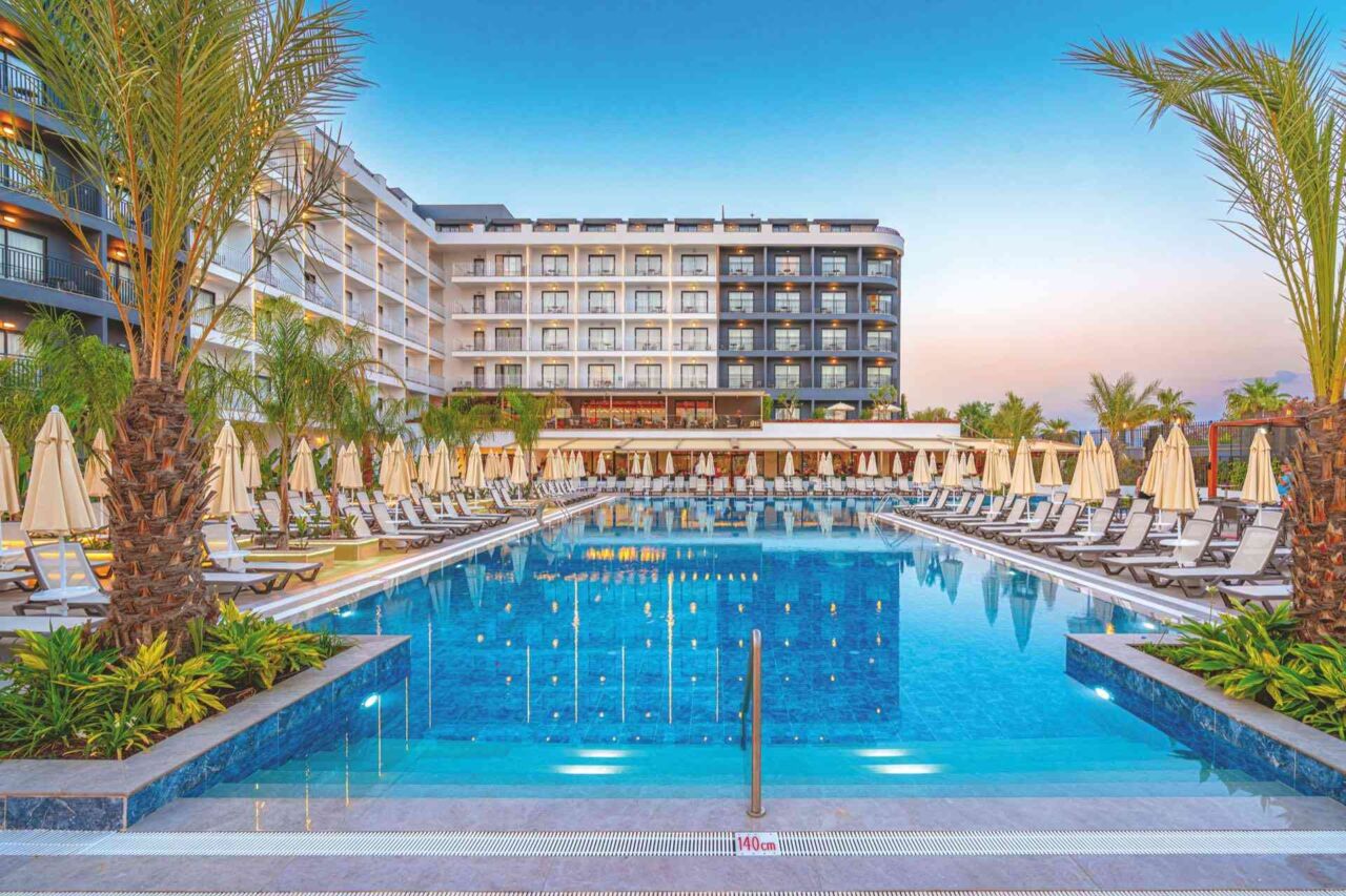 Großer rechteckiger Hotelpool mit Liegestühlen und Sonnenschirmen an beiden Seiten, umgeben von Palmen und einem mehrstöckigen Hotelgebäude im Hintergrund