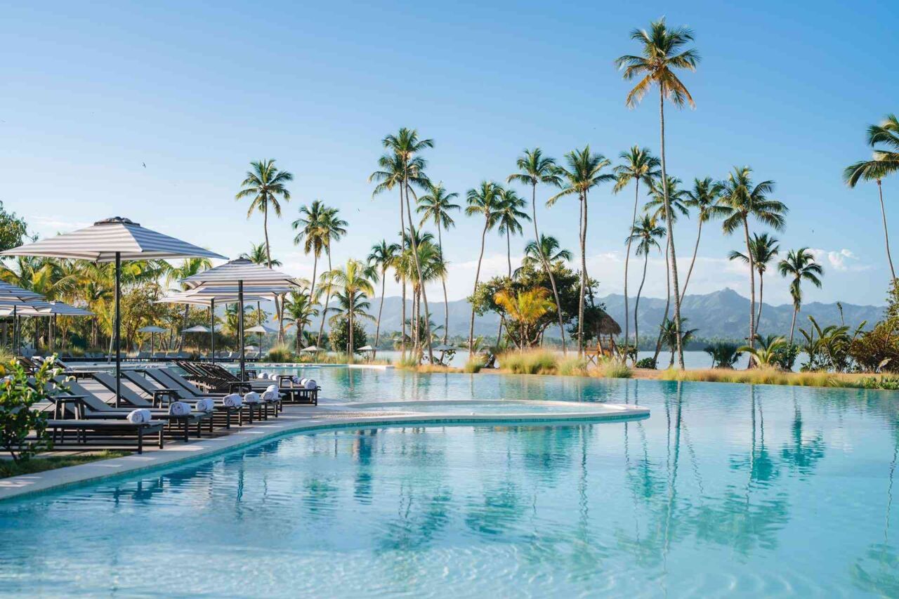 Pool mit Liegestühlen und Sonnenschirmen am Rand, im Hintergrund hohe Palmen und Berge unter blauem Himmel