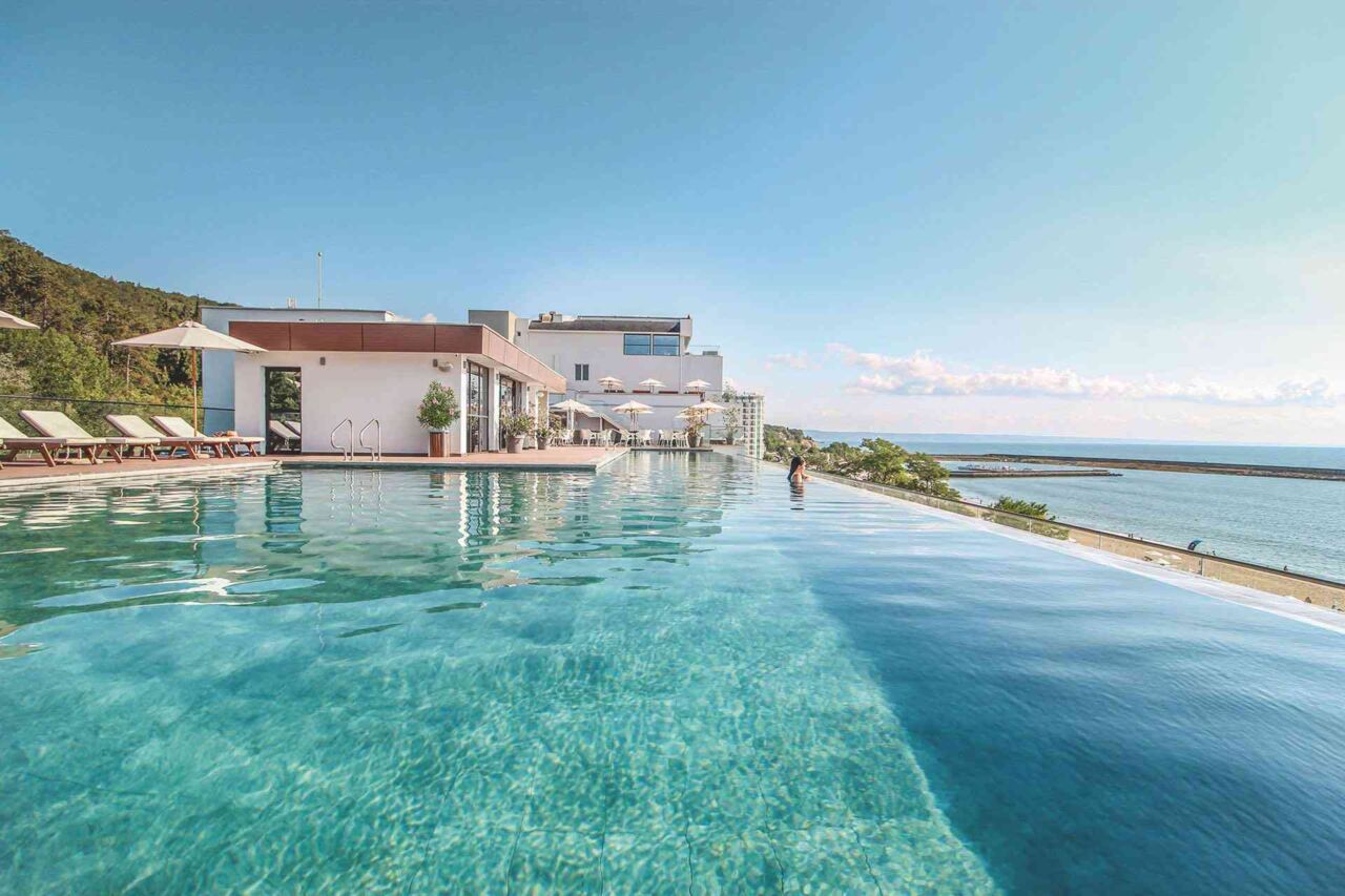 Infinity-Pool mit Liegestühlen und Sonnenschirmen neben modernem Gebäude mit Meerblick